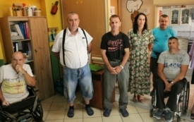 Na pierwszym planie kilkoro stojących dorosłych. W tle ż&oacute;łte ściany a na nich kolorowe dekoracje.