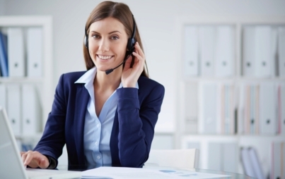 Kobieta siedząca przy biurku. Jedną dłonią trzyma słuchawki, które ma na głowie. Drugą dłoń położoną ma na laptopie. W tle segregatory i meble biurowe.