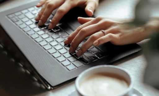 Dłonie kobiety na klawiaturze laptopa. Obok filiżanka kawy.